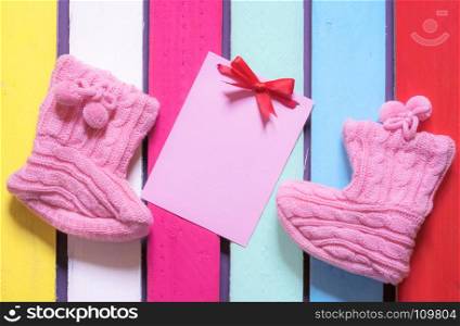 Congratulations on childbirth concept with a message card tied with a red bow, surrounded by cute knitted bootees, on a colorful wooden background.