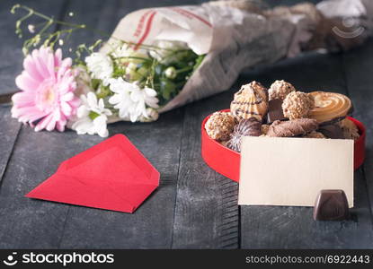 Congratulations theme image with a blank paper note leaned against a box full of sweets, surrounded by a red envelope and a bouquet of flowers, on a wooden table.