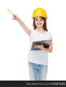 construction and people concept - smiling little girl in protective helmet with clipboard pointing finger at something
