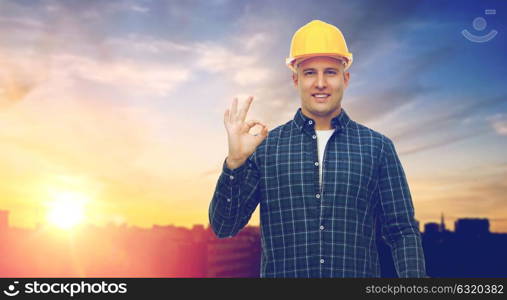 construction, building and gesture concept - smiling male builder or manual worker in helmet showing ok hand sign over city background. smiling builder in helmet showing ok hand sign