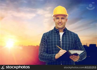 construction, building and people concept - smiling male builder or manual worker in yellow hard hat with clipboard taking notes over city background. male builder in yellow hard hat with clipboard