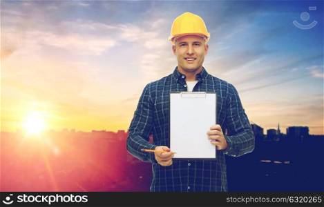 construction, building and people concept - smiling male builder or manual worker in yellow hard hat with clipboard taking notes over city background. male builder in yellow hard hat with clipboard