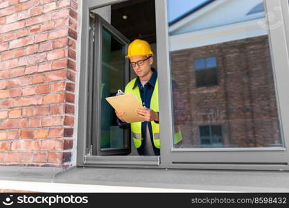 construction busi≠ss and building concept - ma≤builder in helmet and safety west with clipboard and pencil looking out window. ma≤builder with clipboard looking out window