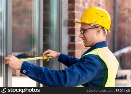 construction business and building concept - happy smiling male builder with ruler measuring window. male builder with ruler measuring window