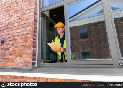 construction business and building concept - male builder in helmet and safety west with clipboard and pencil looking out window. male builder with clipboard looking out window