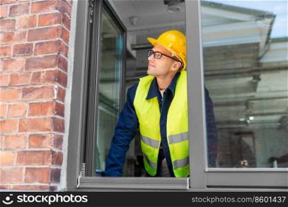 construction business and building concept - male builder in helmet and safety west looking out window. male builder in helmet looking out window