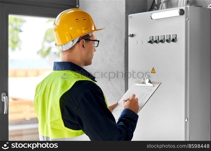 construction business and building concept - male electrician or worker in helmet and safety west with papers on clipboard and pencil at electric board. electrician with clipboard at electric board