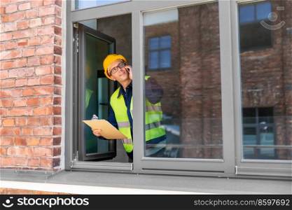 construction business and building concept - smiling male builder in helmet and safety west with clipboard calling on smartphone and looking out window. builder with clipboard and calling on smartphone