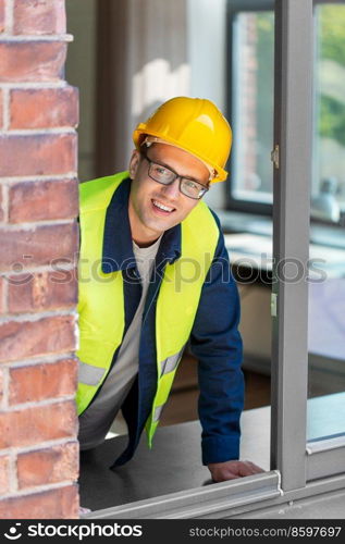 construction business and building concept - smiling male builder in helmet and safety west looking out window. smiling male builder in helmet looking out window