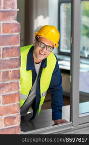 construction business and building concept - smiling male builder in helmet and safety west looking out window. smiling male builder in helmet looking out window