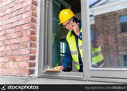 construction business and building concept - smiling male builder in helmet and safety west with clipboard calling on smartphone at window. builder with clipboard and calling on smartphone