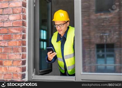 construction business and building concept - smiling male builder in helmet and safety west with smartphone at open window. happy male builder with smartphone at open window
