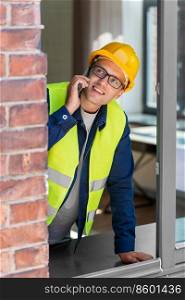construction business and building concept - smiling male builder in helmet and safety west calling on smartphone and looking out window. builder looking out window and calling on phone