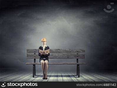 Construction industry. Young woman in hardhat sitting on bench with book