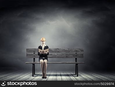 Construction industry. Young woman in hardhat sitting on bench with book