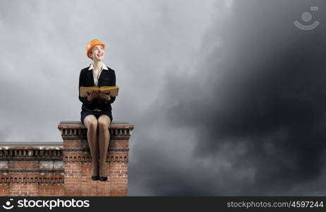 Construction industry. Young woman in hardhat sitting on top of building with book