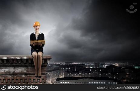 Construction industry. Young woman in hardhat sitting on top of building with book