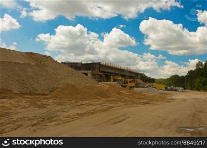 Construction site of a highway leading through the forest with visible sand, gravel and construction machinery under blue sky with white clouds. Poland in summer. Horizontal view.