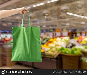consumerism and eco friendly concept - hand holding reusable canvas bag for food shopping over supermarket on background. hand holding reusable canvas bag for food shopping
