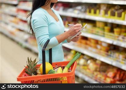 consumerism and people concept - woman with notebook and shopping basket buying food at grocery store or supermarket. woman with food in shopping basket at supermarket