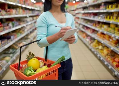 consumerism and people concept - woman with notebook and shopping basket buying food at grocery store or supermarket. woman with food in shopping basket at supermarket