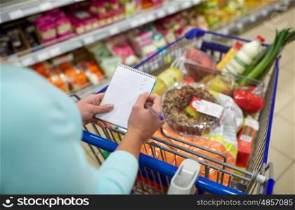 consumerism and people concept - woman with notebook and shopping cart or trolley buying food at grocery store or supermarket. woman with food in shopping cart at supermarket