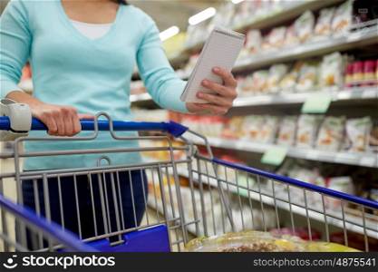consumerism and people concept - woman with notebook and shopping cart or trolley buying food at grocery store or supermarket. woman with food in shopping cart at supermarket