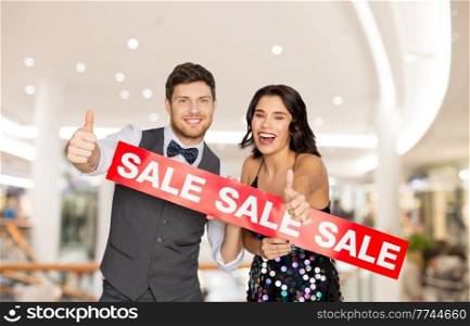 consumerism, people and fashion concept - happy couple with red sale sign showing thumbs up over shopping mall background. happy couple with red sale sign showing thumbs up