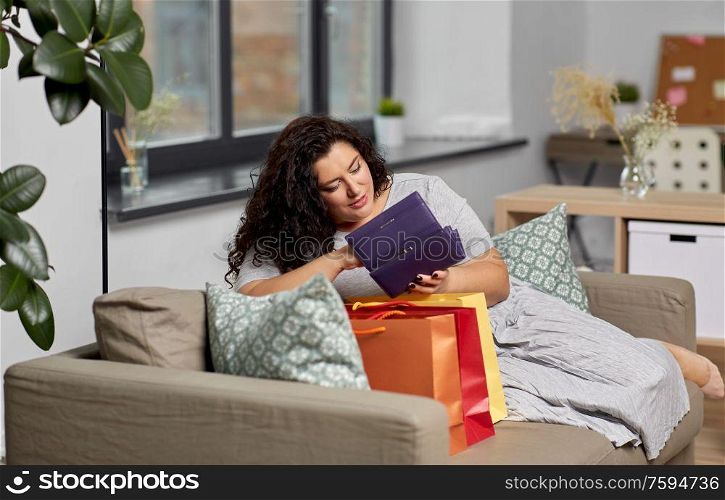 consumerism, people and sale concept - young woman with shopping bags and wallet sitting on sofa at home. woman with shopping bags and wallet at home