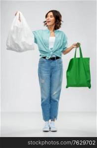 consumerism, sustainability and eco living concept - portrait of happy smiling young woman in turquoise shirt and jeans with plastic and green reusable canvas bag for food shopping on grey background. woman with plastic and reusable shopping bag