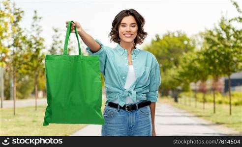 consumerism, sustainability and people concept - portrait of happy smiling young woman in turquoise shirt and jeans with green reusable canvas bag for food shopping over city street background. woman with reusable canvas bag for food shopping