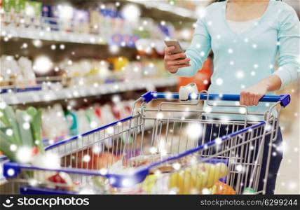 consumerism, technology and people concept - female customer with smartphone and shopping cart or trolley buying food at grocery store or supermarket over snow. woman with smartphone buying food at supermarket