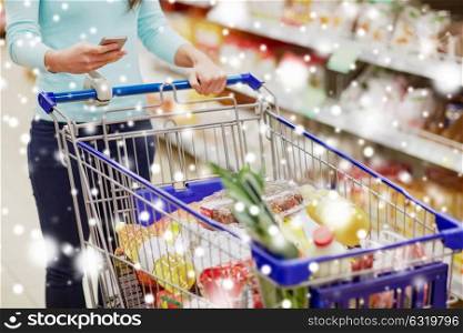 consumerism, technology and people concept - female customer with smartphone and shopping cart or trolley buying food at grocery store or supermarket over snow. customer with food in shopping cart at supermarket