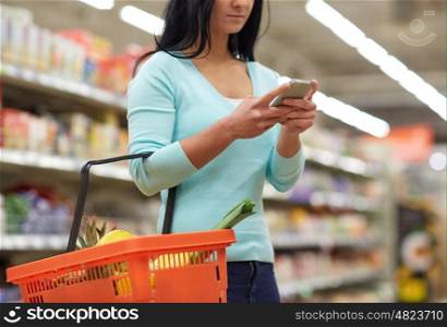 consumerism, technology and people concept - woman with smartphone and shopping basket buying food at grocery store or supermarket