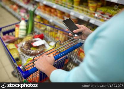 consumerism, technology and people concept - woman with smartphone and shopping cart or trolley buying food at grocery store or supermarket