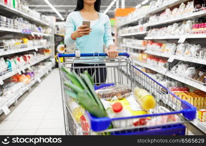 consumerism, technology and people concept - woman with smartphone and shopping cart or trolley buying food at grocery store or supermarket. woman with smartphone buying food at supermarket