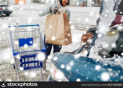 consumerism, transportation and people concept - female customer loading food from shopping cart to car trunk at winter parking over snow. customer loading food from shopping cart to car