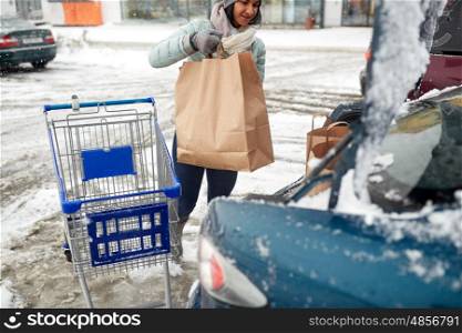 consumerism, transportation and people concept - woman loading food from shopping cart to car trunk at winter parking