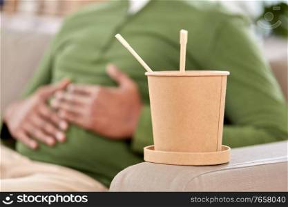 consumption and people concept - cup of takeaway food in front of overeaten man at home. cup of takeaway food over man at home