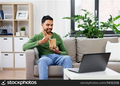 consumption and people concept - smiling indian man eating takeaway food and watching something on laptop computer at home. indian man with laptop eating takeout food at home