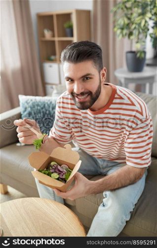 consumption, delivery and people concept - smiling man with fork and knife eating takeaway food at home. smiling man eating takeaway food at home