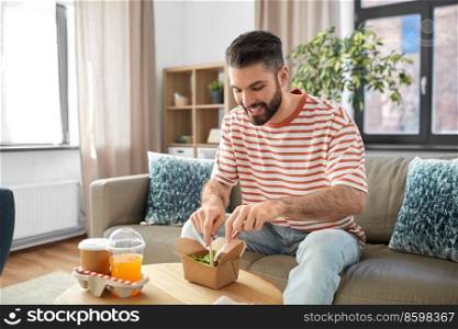 consumption, delivery and people concept - smiling man with fork and knife eating takeaway food at home. smiling man eating takeaway food at home