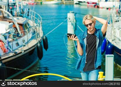 Contacts, technology concept. Hipster young blonde man on vacations holding his smartphone. Outdoor in marina, sunny day. Blonde man outside using his smartphone
