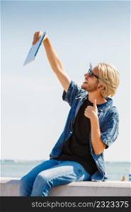 Contacts, technology, self-esteem concept. Young blonde man on vacations using his tablet to take cool selfie shot, showing thumb up gesture. Hipster man outside taking selfie with tablet
