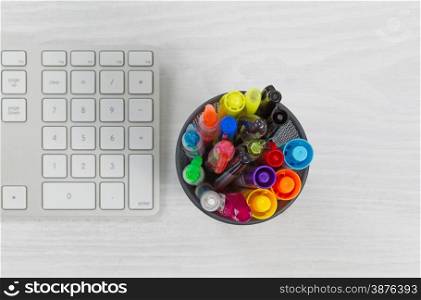 Container of pens and markers, focus top of container, and computer keyboard on rustic white desktop.