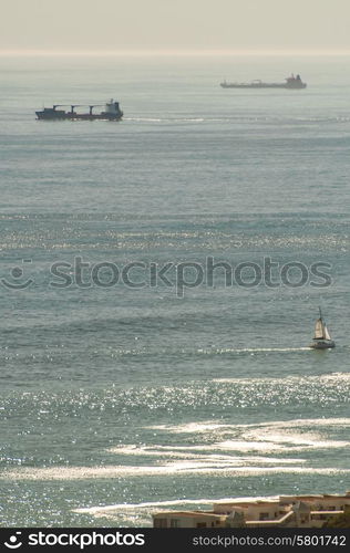 Container ships travel in the hazy distance, near the horizon, on the cold Atlantic ocean near Cape Point, while closer to the apartment buildings visible below, a small yacht sails in the opposite direction.