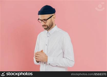 Contemplative unshaven man keeps gaze down, wears white shirt, hat and glasses, being deep in thoughts, tries to make solution, thinks how develop his business, isolated over pink background