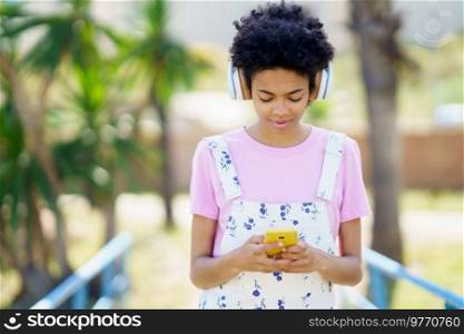 Content African American female listening to song in wireless headphones while scrolling cellphone on street against blurred background on summer day. Black woman listening to music while browsing smartphone