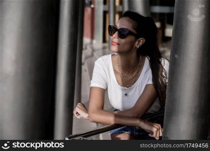 Content beautiful woman in sunglasses and shorts leaning on metal handrail while standing at terrace on sunny day. Relaxed woman in sunglasses resting while leaning on street handrail