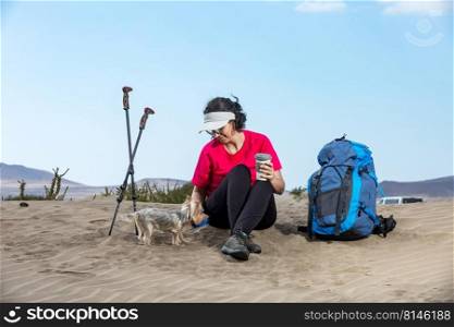 Content female backpacker sitting with Yorkshire Terrier dog on sandy Famara beach and drinking beverage while taking break during hike. Traveling woman with dog on seashore